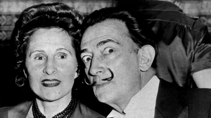 Schwarzweiß-Foto von Gala und Dalí bei einem Essen. Dalí beugt sich zu ihr hin und verdreht die Augen, sie lächelt in die Kamera. | Bildquelle: dpa Schwarzweiß-Foto von Gala und Dalí bei einem Essen. Dalí beugt sich zu ihr hin und verdreht die Augen, sie lächelt in die Kamera.