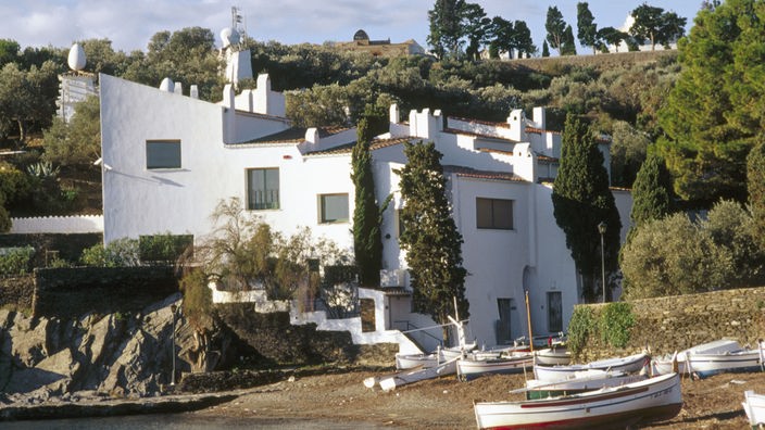 Blick auf ein weißes, verschachteltes Haus direkt an einer kleinen Bucht. An dem schmalen Sandstrand liegen ein paar Boote.