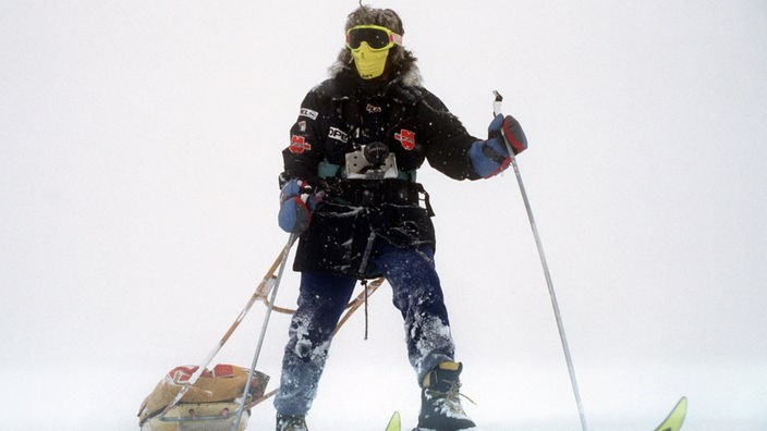Das Foto zeigt Reinhold Messner dick vermummt mit Schutzmaske, Skibrille und Skianzug im Schnee. Er geht auf Skiern und zieht einen kleinen Lastenschlitten hinter sich her.