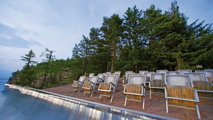 Das Foto zeigt mehrere Reihen von Stühlen aus Stahl und Holz auf einer sandigen Fläche unter freiem Himmel. Dahinter sind hohe grüne Bäume und ein begrünter Felsen zu sehen.