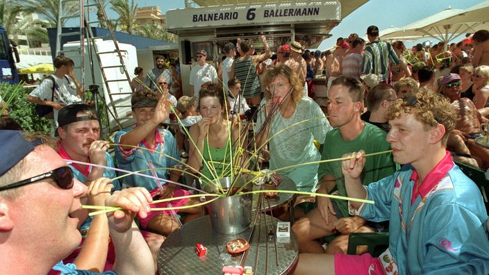 Sieben Touristen trinken mit Strohhalmen aus einem gemeinsamen Topf, der auf einem runden Tisch steht. Im Hintergrund mehr Menschen und die Strandbude mit der Aufschrift 'Ballermann - 6 - Balneario'.