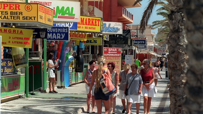Einkaufsstraße in El Arenal. Rechts gehen Passanten, links sieht man die Schilder der Läden wie 'Kalter Sekt', 'Sangria' oder 'Souvenirs'.