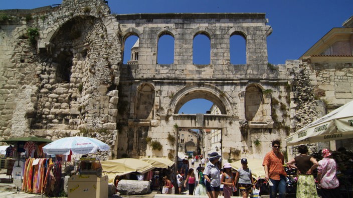 Menschen und Marktstände vor den Ruinen des Eingangsportals eines römischen Palastes