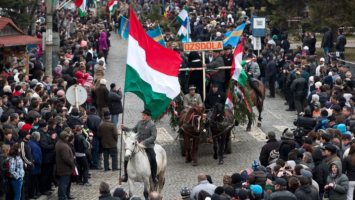 Auf einer Straßenparade hält ein Reiter eine große ungarische Flagge. | Bildquelle: picture alliance / AP Auf einer Straßenparade hält ein Reiter eine große ungarische Flagge.