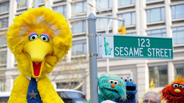 Bibo und andere Puppen neben einem Straßenschild mit der Aufschrift "123 Sesame Street"
