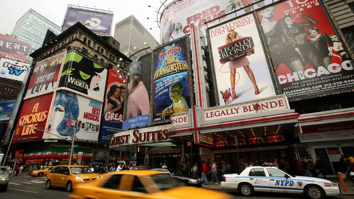 New Yorker Broadway am Tage. Ein Musicaltheater mit Reklametafel reiht sich an das nächste. Auf der Straße dichter Taxiverkehr.