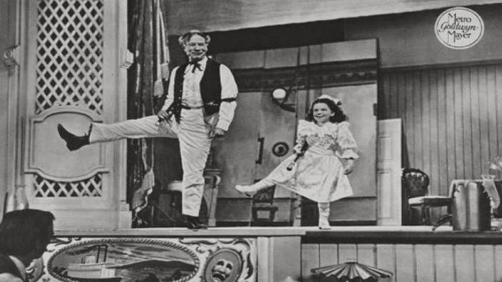 Ein Schwarzweißfoto zeigt eine Szene aus der Verfilmung des Musicals 'Show Boat'. Man sieht einen Mann und ein Mädchen. Beide tanzen auf einer Bühne. Davor ist ein Zuschauer zu sehen.