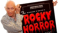 Richard O'Brien hält ein Werbeplakat für die Aufführung der 'Rocky Horror Show' in den Händen.