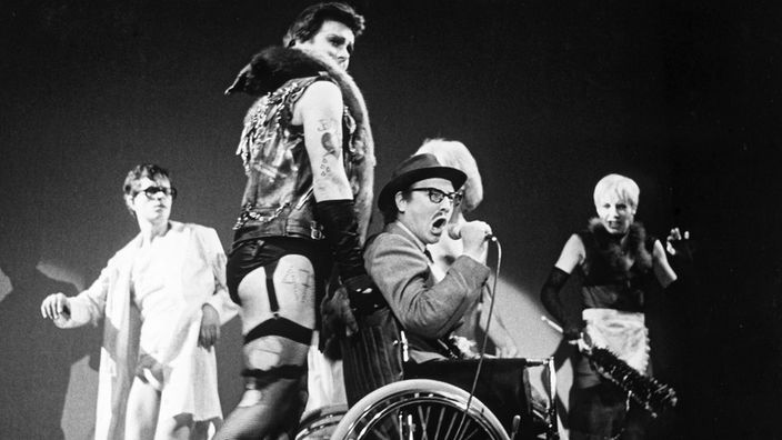 Ein Schwarzweiß-Foto zeigt eine Szene aus dem Musical 'Rocky Horror Show' Zu sehen ist Frank'n'Furter, der einen Rollstuhl vor sich her schiebt.