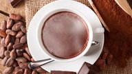 Eine Tasse Kakao (heiße Schokolade) steht zwischen Kakaopulver, Kakaobohnen und Schokoladenstücken