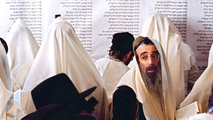 Mehrere Männer mit Gebetsmänteln über dem Kopf stehen vor einer Wand, auf der hebräische Schriftzeichen zu sehen sind.
