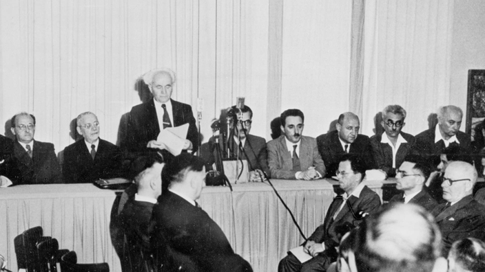 Schwarzweiß-Foto: In einem Raum sitzen mehrere Männer an einem langen Tisch. Einer der Männer steht und hat Papiere in der Hand. An der Wand sind zwei israelische Staatsbanner befestigt.