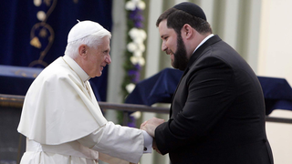 Papst Benedikt in weißer Kleidung schüttelt einem schwarz gekleideten Rabbiner die Hand.