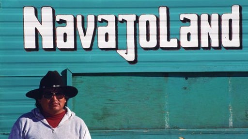 Ein Indianer mit Cowboyhut und Sonnenbrille steht vor einem Schild mit der Aufschrift 'Navajoland'.