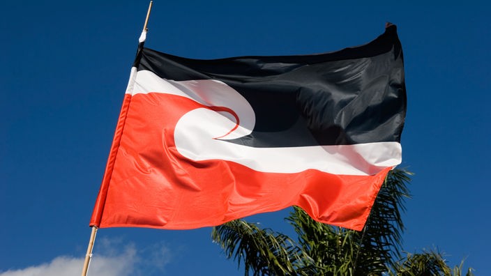 Das Bild zeigt eine Flagge, die im oberen Teil schwarz und im unteren Teil rot ist und in der Mitte eine Art weiße Welle hat, vor blauem Himmel.