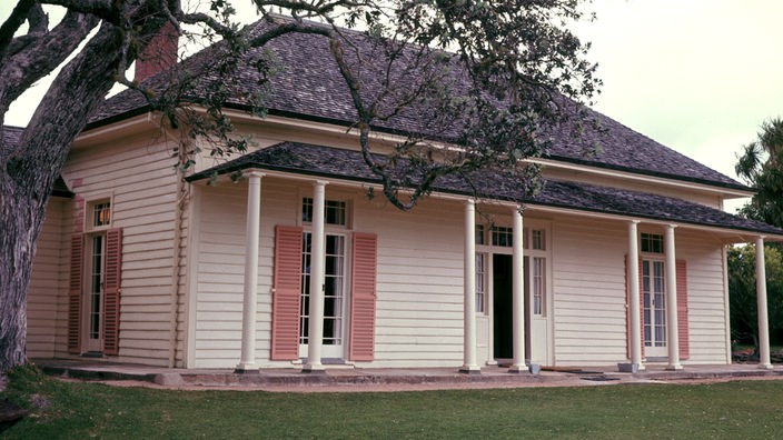 Das Bild zeigt ein relativ kleines Haus aus weißem Holz mit schwarzem Dach. Es hat ein kleines Vordach, das ebenfalls schwarz ist. Die Fensterläden sind lachsfarben. Links neben dem Haus steht ein großer Baum.