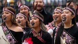 Das Bild zeigt eine Gruppe von Maori-Männern mit traditionellen schwarzen Tätowierungen im Gesicht. Ein Mann im Vordergrund streckt die Zunge heraus und hat die Augen weit aufgerissen. Er hält einen langen Holzstab in der Hand.