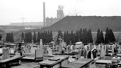 Kohlegeschwärzte Gräber im Vordergrund, im Hintergrund ein heruntergekommenes Kohlekraftwerk in der Wallonie.