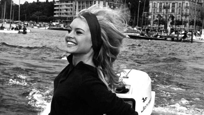 Im Vordergrund eine lachende langhaarige Blondine auf einem Motorboot. Im Hintergrund ein Jachthafen und zwei große Gebäude.