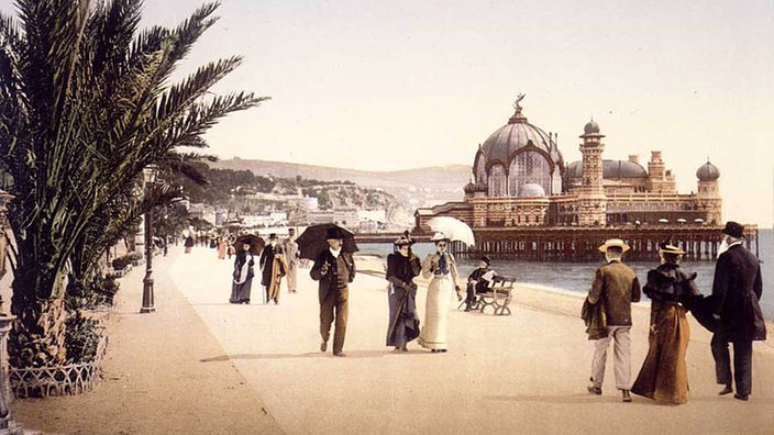Das kolorierte Foto zeigt eine breite Uferpromenade mit flanierenden Menschen. Die Damen tragen Sonnenschirme.