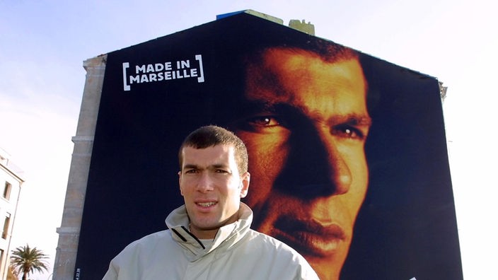 Zinédine Zidane steht vor einer Häuserwand, auf der sein Gesicht abgebildet ist