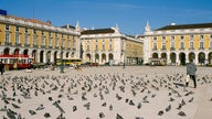 Tauben tummeln sich auf einem großen Platz, der eingerahmt von arkadengeschmückten Gebäuden ist.