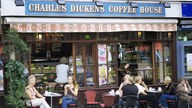 Draußen vor einem Café sitzen Passanten an Bistrotischen bei einem Kaffee oder Tee. Die Türen nach innen sind auf, es ist eine Theke zu sehen, oben steht der Name des Cafés: 'Charles Dickens Coffee Hous