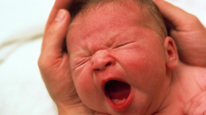 Ein gähnender Säugling, dessen Kopf in den Händen eines Erwachsenen liegt.