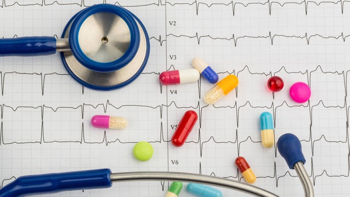 Tabletten und Pillen liegen neben einem Stethoskop auf dem Ausdruck eines EKGs.