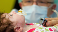 Ein Kleinkind mit Schnulli im Mund wird von einem Arzt mit dem Stethoskop an der Brust abgehört