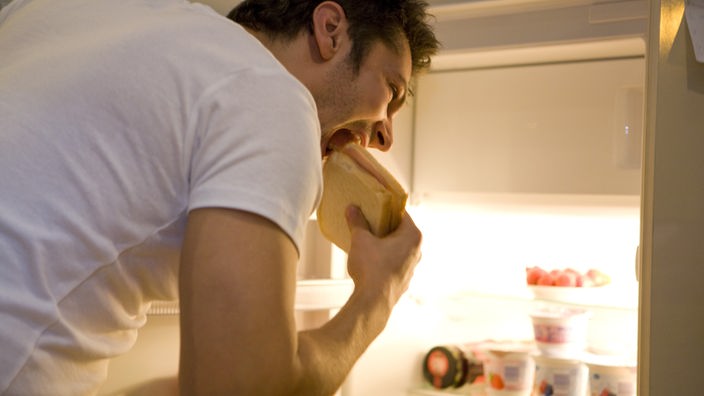 Mann steht am Kühlschrank und isst ein Sandwich