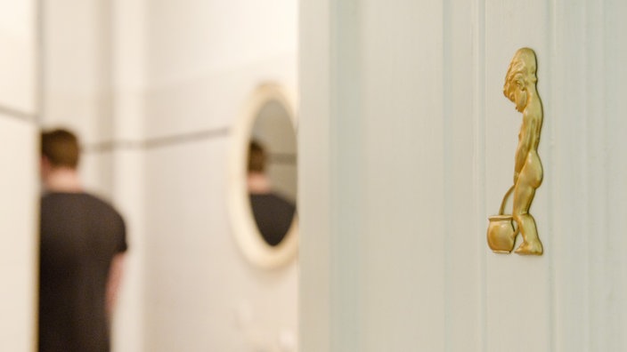 WC-Schild in Form eines Jungen während ein Mann im Hintergrund die Toilette benutzt | Bildquelle: dpa Picture-Alliance/dpa Themendienst/Franziska WC-Schild in Form eines Jungen während ein Mann im Hintergrund die Toilette benutzt