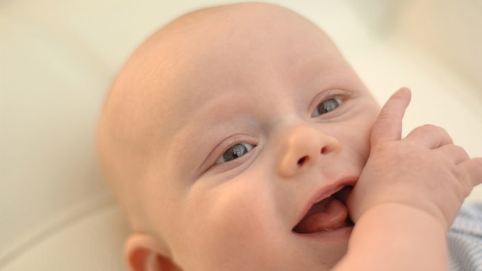 Porträtaufnahme eines Babys, das am Daumen lutscht und in die Kamera lächelt.