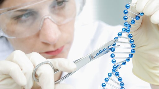 Frau mit Schutzbrille und Laborkleidung hält ein Modell einer DNA-Doppelhelix in der Hand und bearbeitet es mit einer Schere.
