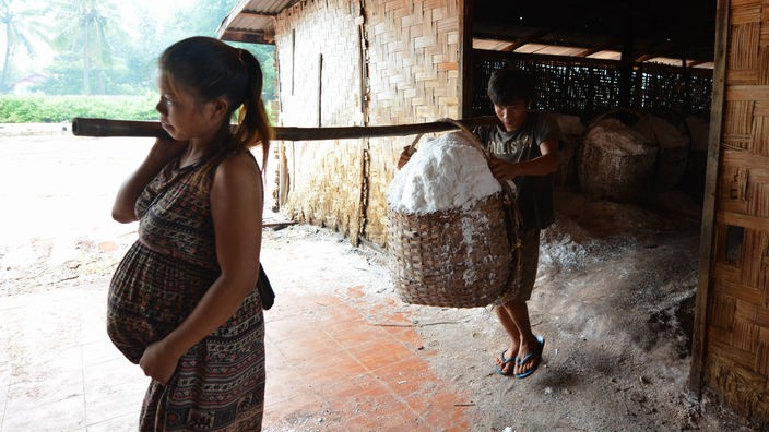Eine schwangere Frau und ejn junger Mann tragen einen Salzkorb, der an ener Holzstange hängt
