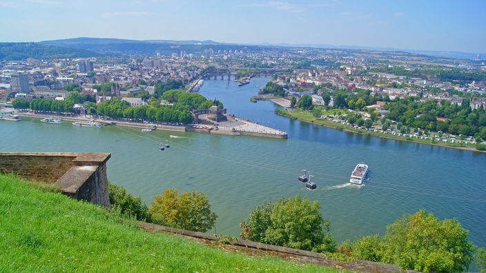 Im Vordergrund ist der Lauf des Rheins zu sehen, der eine leicht bräunliche Färbung hat. Am sogenannten Deutschen Eck, mit dem Reiterstandbild von Kaiser Wilhelm I., fließt die Mosel in den Rhein. Sie hat eine bläuliche Wasserfärbung.