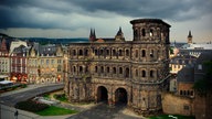 Eine Stadtansicht von Trier am frühen Abend. Im Vordergrund die römische Porta Nigra. Am Himmel ziehen dunkle Gewitterwolken auf.