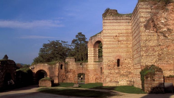 Steil aufragende Ruinenfassade der römischen Kaiserthermen. In den aus rotem Ziegel gemauerten Wänden sind Rundfensterbögen zu sehen. Die antike Kulisse wird von der sommerlichen Abendsonne beschienen.