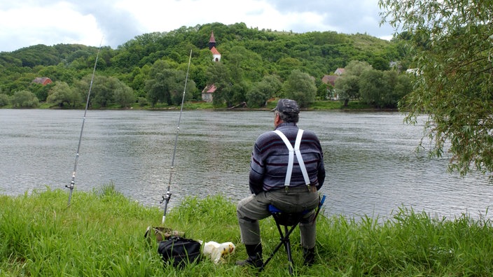 Ein Angler sitzt im Nationalpark Unteres Odertal an der Oder. Neben ihm stehen zwei Angeln und sein Fahrrad. Am gegenüberliegenden Flussufer steht eine Kapelle. | Bildquelle: dpa/Patrick Pleul Ein Angler sitzt im Nationalpark Unteres Odertal an der Oder. Neben ihm stehen zwei Angeln und sein Fahrrad. Am gegenüberliegenden Flussufer steht eine Kapelle.