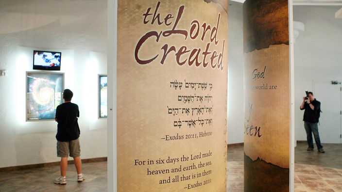 Das Bild zeigt einen Museumsraum. Im Vordergrund sind zwei Tafeln zu sehen. Auf der einen steht in großen Buchstaben 'the Lord Created'.