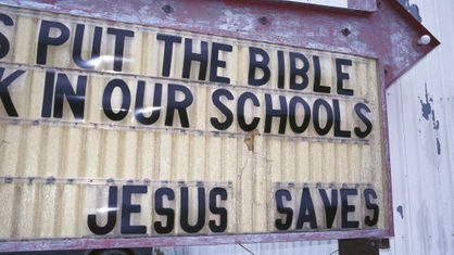 Das Bild zeigt ein Schild, das vor einem Wellblechgebäude steht, darauf in Großbuchstaben der Text 'Put the bible in our schools - Jesus saves.'