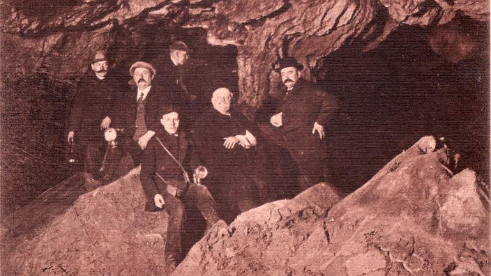 Sechs gut bekleidete Männer stehen in einer Höhle und posieren für ein Foto
