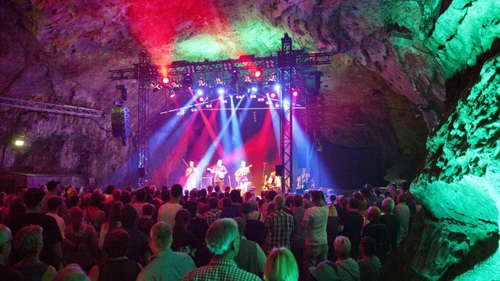 Zuschauer stehen vor einer bunt beleuchteten Bühne in einer Höhle