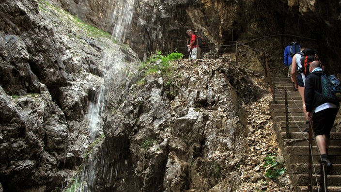 Wasser stürzt über Felsen in die Tiefe. Am rechten Bildrand sieht man Wanderer auf einer Felsentreppe.