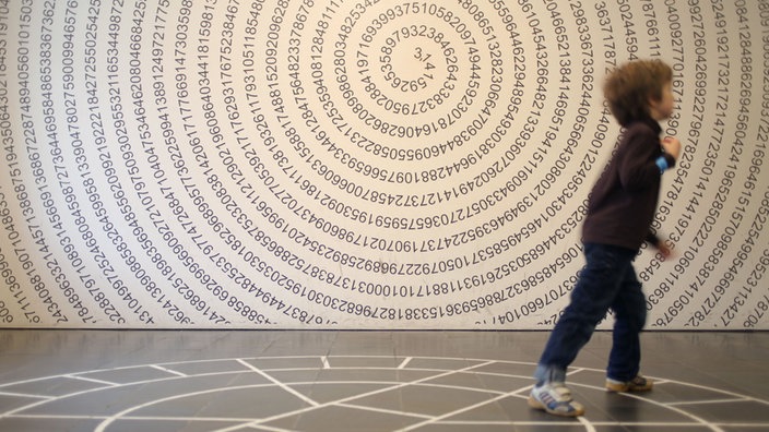 Fotografie eines Kindes vor einer Wand, auf der die Stellen der Zahl Pi spiralförmig angeordnet sind | Bildquelle: dpa/Emily Wabitsch Fotografie eines Kindes vor einer Wand, auf der die Stellen der Zahl Pi spiralförmig angeordnet sind