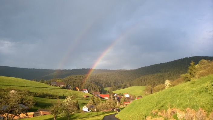 Ein doppeltes Regenbogenstück über einigen Häusern zwischen Wiesen und Wäldern.