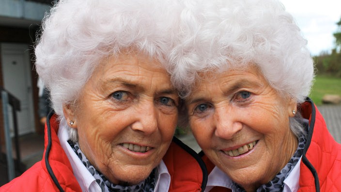 Zwei ältere eineiige Zwillingsfrauen mit weißen Haaren und roter Jacke.