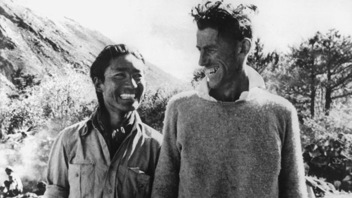 Schwarzweiß-Bild: Zwei Männer stehen nebeneinander und grinsen sich an.