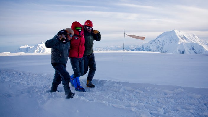 Ein Mann wird von zwei anderen Männern auf einem schneebedeckten Gipfel unter die Arme genommen