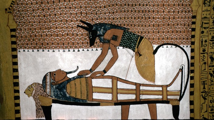 Die ägyptische Wandmalerei zeigt den Gott Anubis, der einen Hundekopf hat, wie er einen Toten bandagiert.